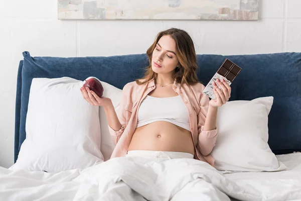 Красива вагітна жінка лежить в ліжку з шоколадним баром і яблуком — стокове фото