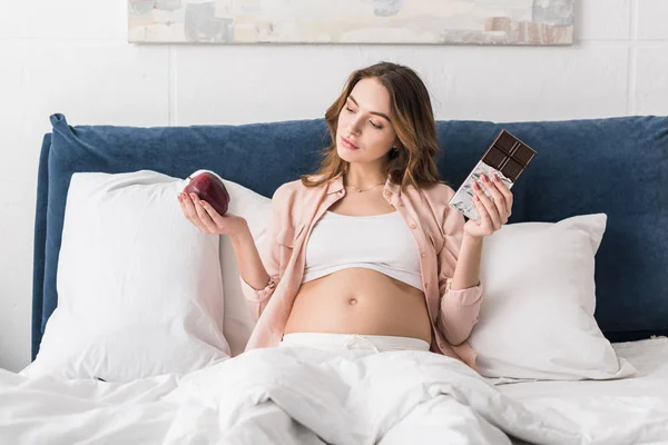Beautiful pregnant woman lying in bed with chocolate bar and apple — Stock Photo