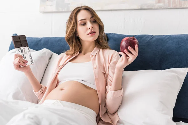 Pregnant woman holding red apple and chocolate bar — Stock Photo
