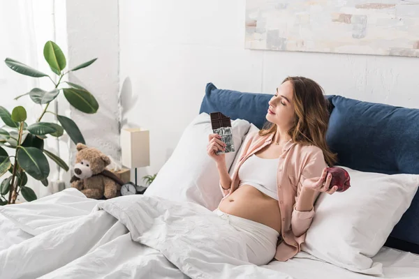 Pregnant woman lying in bed with chocolate bar and red apple — Stock Photo