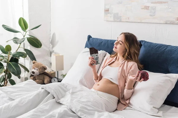 Mujer embarazada acostada en la cama con barra de chocolate y manzana roja - foto de stock