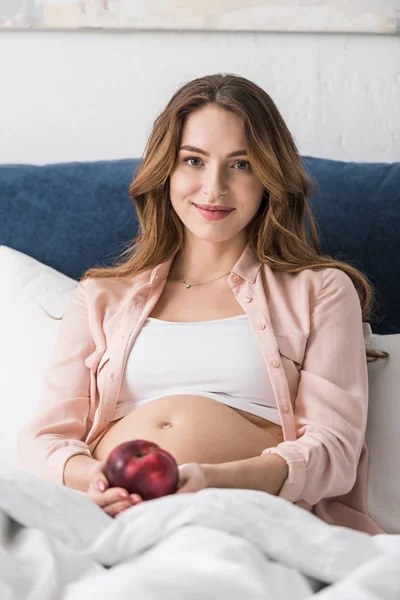 Щаслива вагітна жінка лежить в ліжку з яблуком і дивиться на камеру — стокове фото