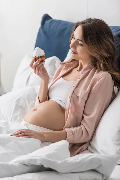 Beautiful pregnant woman lying in bed and holding cupcake — Stock Photo