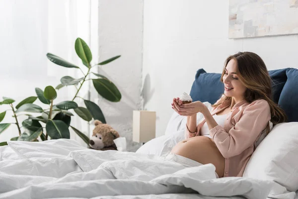Pretty pregnant woman lying in bed and holding cupcake — Stock Photo