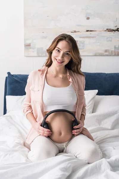 Felice donna incinta seduta sul letto con le cuffie sulla pancia — Foto stock