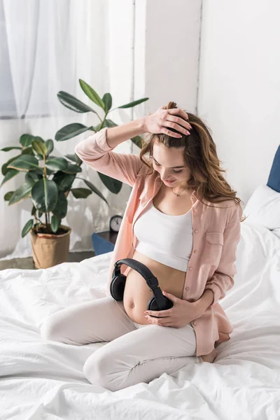 Mujer embarazada sentada en la cama y sosteniendo auriculares en el vientre - foto de stock