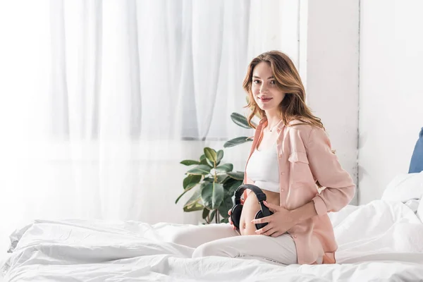 Красива вагітна жінка сидить на ліжку з навушниками на животі — стокове фото