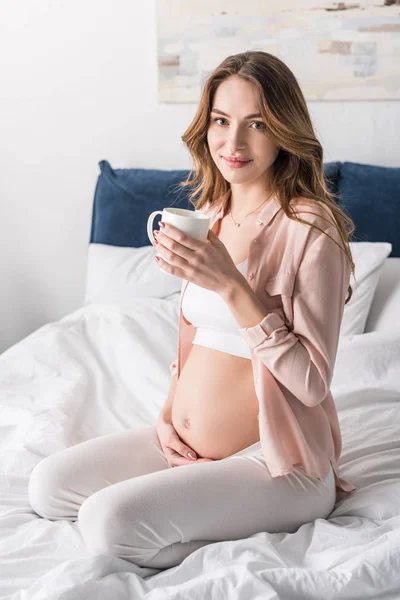 Schöne schwangere Frau sitzt mit Kaffeetasse im Bett — Stockfoto