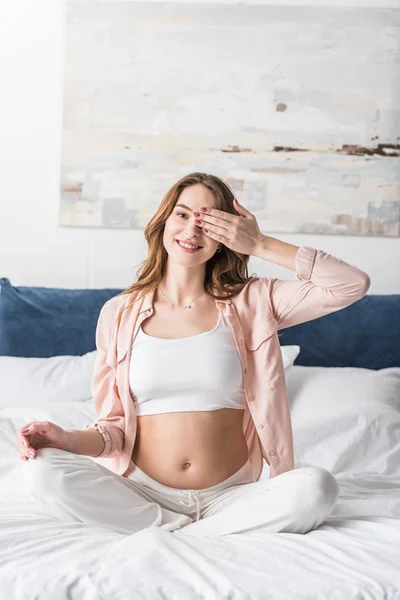 Блаженна вагітна жінка сидить на ліжку і прикриває око рукою — стокове фото