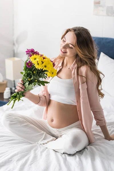 Smiling pregnant woman sitting on bed with flowers — Stock Photo