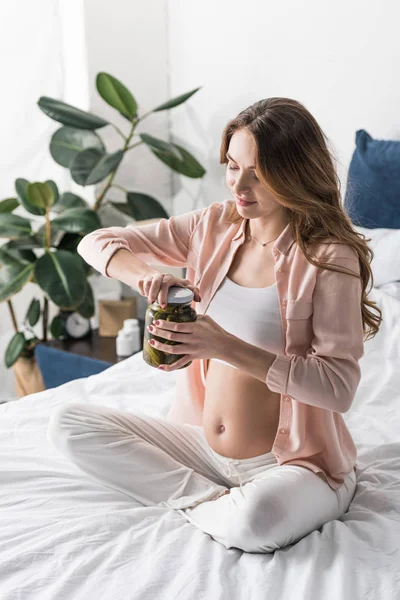 Femme enceinte assise sur le lit et ouvrant le pot avec des cornichons — Photo de stock