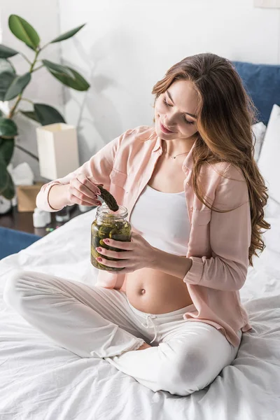 Beautiful pregnant woman eating salted cucumbers on bed — Stock Photo