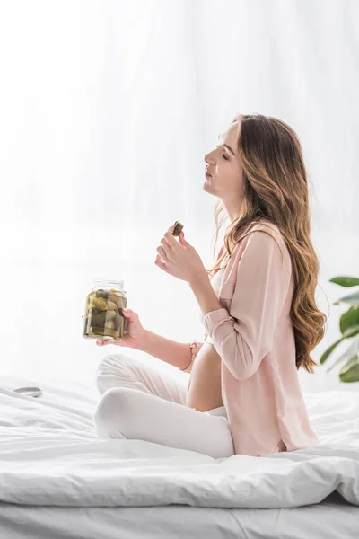 Side view of pregnant woman sitting on bed and eating pickles — Stock Photo