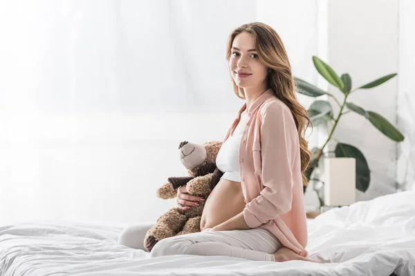Чарівна вагітна жінка сидить на ліжку з плюшевим ведмедем — Stock Photo