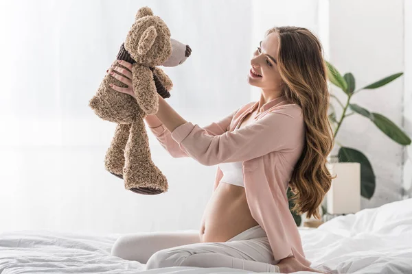 Femme enceinte aux cheveux longs assise sur le lit et regardant un ours jouet — Photo de stock