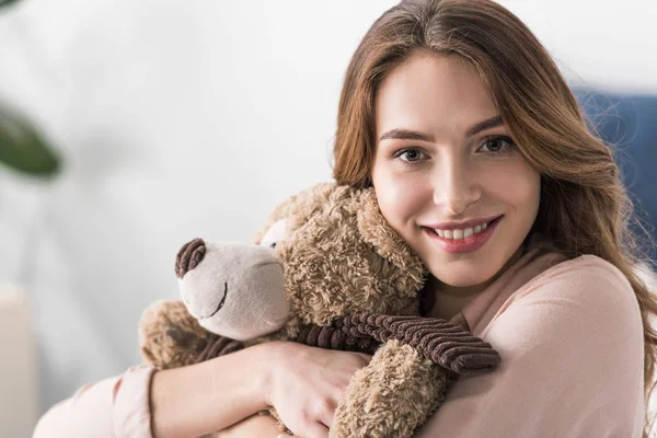 Mujer sonriente abrazando oso de juguete y mirando a la cámara - foto de stock