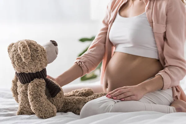 Vista cortada da mulher grávida sentada na cama com urso de brinquedo — Fotografia de Stock