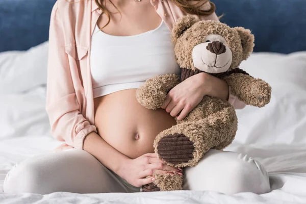 Cropped view of pregnant woman with teddy bear sitting on bed — Stock Photo