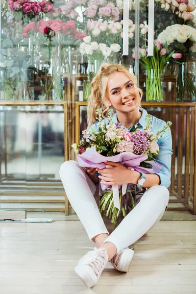 Hermosa mujer sonriente sosteniendo ramo de flores, mirando a la cámara y sentado frente a la tienda de flores - foto de stock