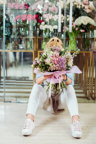 Mujer sosteniendo ramo de flores en frente de la cara mientras está sentado cerca de la tienda de flores - foto de stock