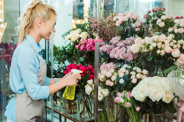 Жіночий флорист в фартуху розбризкує квіти з пляшкою розпилення в квітковому магазині — стокове фото