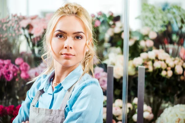 Enfoque selectivo de hermosa florista femenina en delantal mirando a la cámara con floristería en el fondo - foto de stock