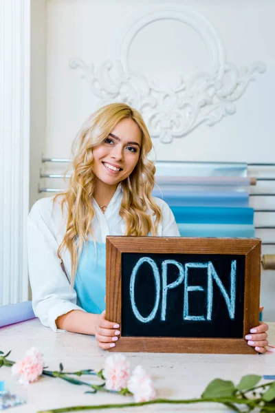 Beautiful smiling female flower shop owner looking at camera and holding chalkboard with 'open' lettering — Stock Photo