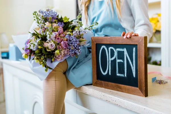 Vista recortada de la propietaria de la floristería femenina sosteniendo pizarra con letras 'abiertas' y colorido ramo - foto de stock