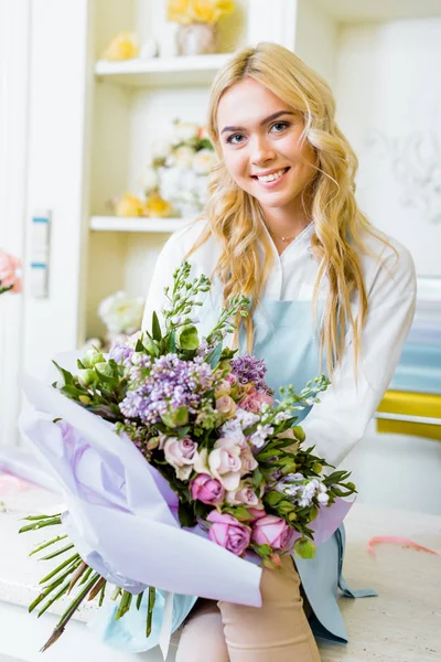 Belle propriétaire de magasin de fleurs femelle regardant la caméra, assis sur le comptoir et tenant bouquet — Photo de stock