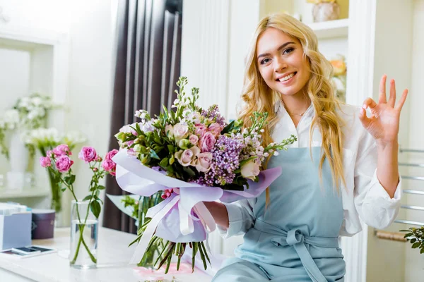 Bonito sorridente fêmea flor loja proprietário sittng no balcão e segurando buquê enquanto mostrando sinal ok — Fotografia de Stock