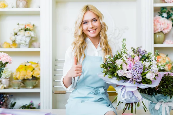 Schöne lächelnde Blumenverkäuferin sitzt auf der Theke und hält Strauß, während sie Daumen nach oben Zeichen zeigt — Stockfoto