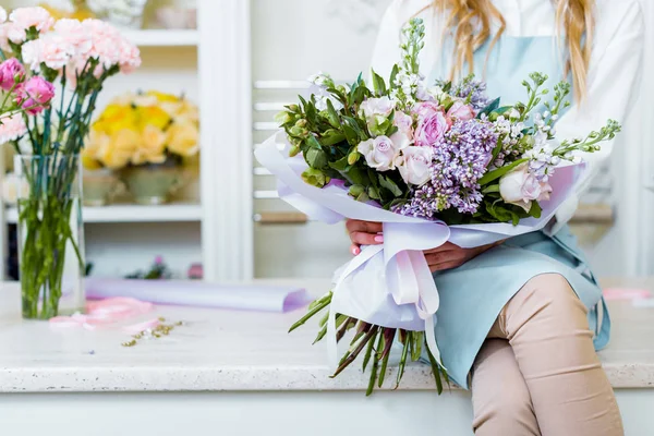 Обрезанный вид женщины-флориста, сидящей на прилавке и держащей букет с сиренью и розами в цветочном магазине — стоковое фото