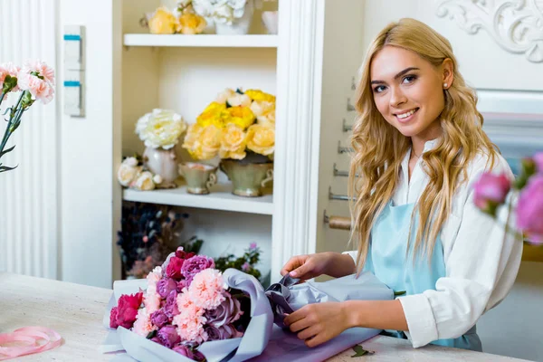 Hermosa florista femenina sonriente que arregla y envuelve el ramo en la florería - foto de stock