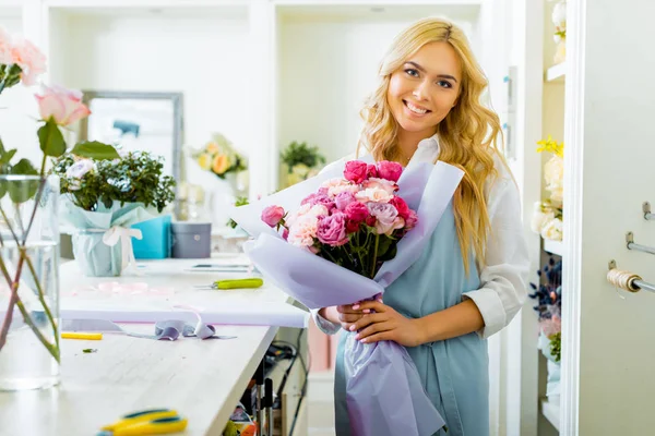 Beautiful female florist looking at camera and holding bouquet in flower shop — Stock Photo