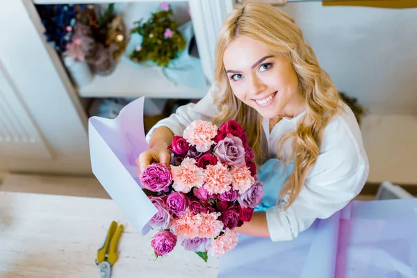 Bela florista feminina olhando para a câmera enquanto segurando buquê com rosas e cravos na loja de flores — Fotografia de Stock