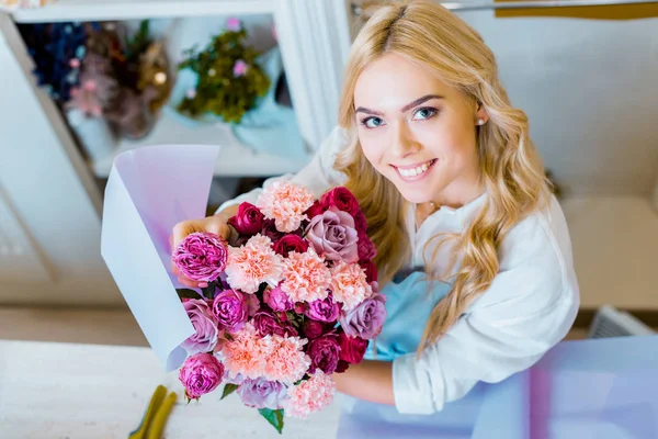 Schöne Floristin blickt in die Kamera, während sie einen Strauß mit Rosen und Nelken im Blumenladen hält — Stockfoto