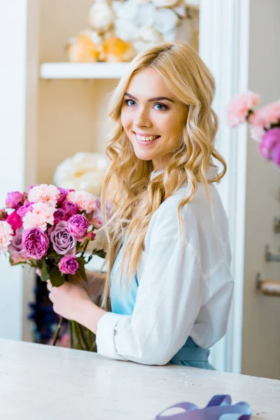 Beautiful female florist holding bouquet with roses and carnations in flower shop and looking away — Stock Photo