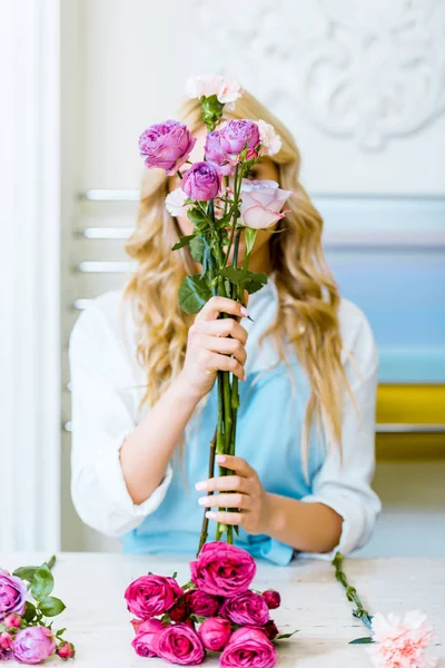 Florista feminina em avental segurando rosas na frente do rosto enquanto organiza buquê na loja de flores — Fotografia de Stock