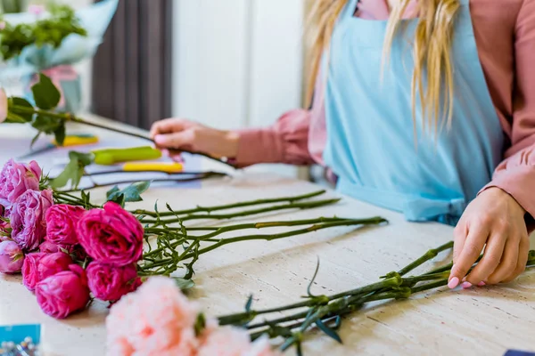 Vista cortada de florista feminino sentado à mesa com rosas rosa e cravos enquanto organiza buquê — Fotografia de Stock