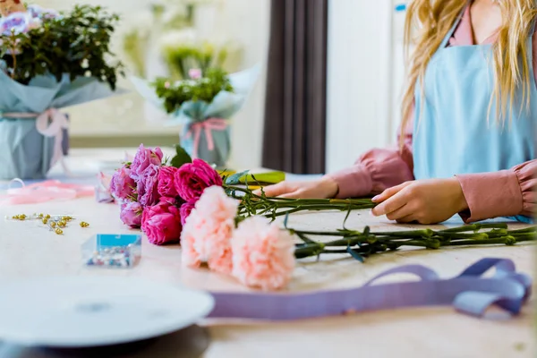 Cropped view of female florist sitting at table with pink roses and carnations while arranging bouquet — Stock Photo