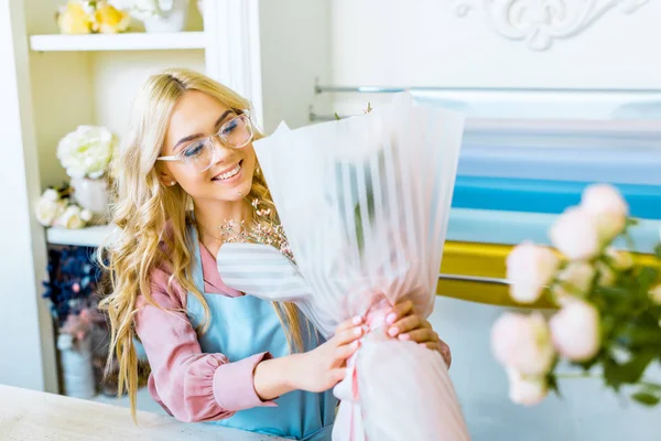 Hermosa rubia florista femenina en gafas sonriendo y sosteniendo ramo en floristería - foto de stock