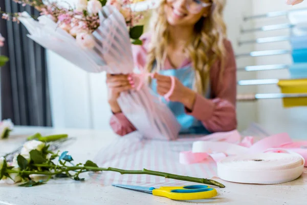 Partial view of female florist arranging bouquet in flower shop with ribbon and scissors on foreground — Stock Photo