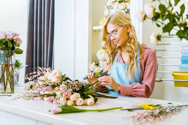 Hermosa florista femenina en gafas que arreglan ramo y corte de rosas con podadora en floristería - foto de stock