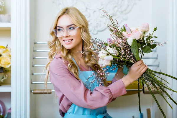 Красивая женщина-флористка в очках держит букет в цветочном магазине и смотрит в сторону — стоковое фото