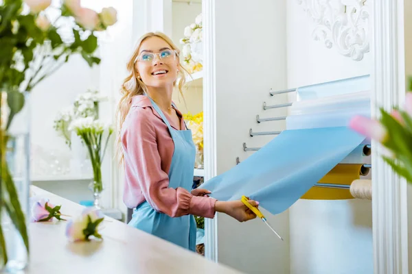 Beautiful smiling female florist with wrapping paper and scissors in flower shop — Stock Photo