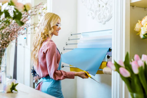 Hermosa florista femenina sonriente con papel de envolver y tijeras en la tienda de flores - foto de stock