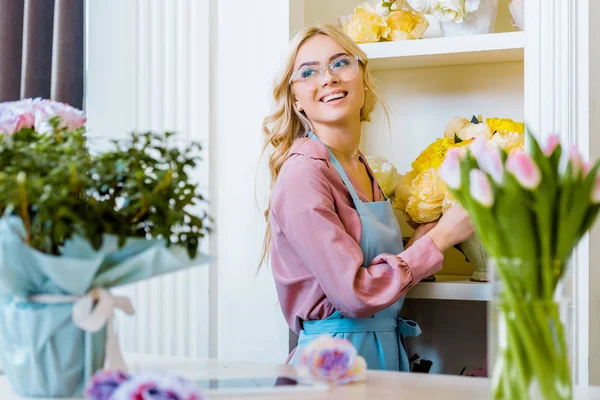 Beautiful happy female florist in glasses holding yellow bouquet in flower shop — Stock Photo