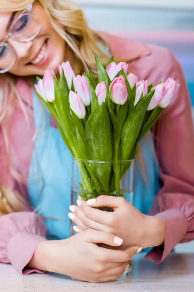 Hermosa florista femenina en gafas sonriendo y sosteniendo jarrón con tulipanes rosados en floristería - foto de stock
