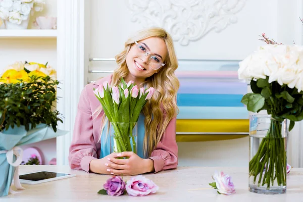 Hermosa florista femenina mirando a la cámara y sosteniendo florero con tulipanes rosados en la tienda de flores - foto de stock