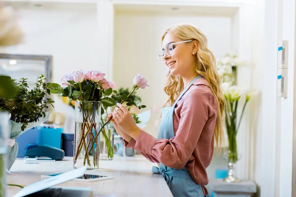 Hermosa floristería femenina sonriente poner rosa rosa en ramo en la tienda de flores - foto de stock